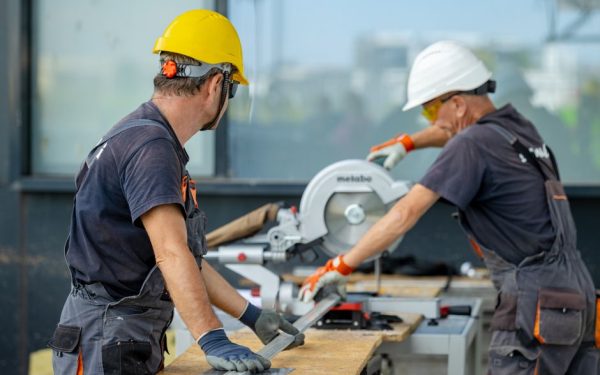 Two construction workers cutting a metal beam with a circular saw, wearing safety gear on site.