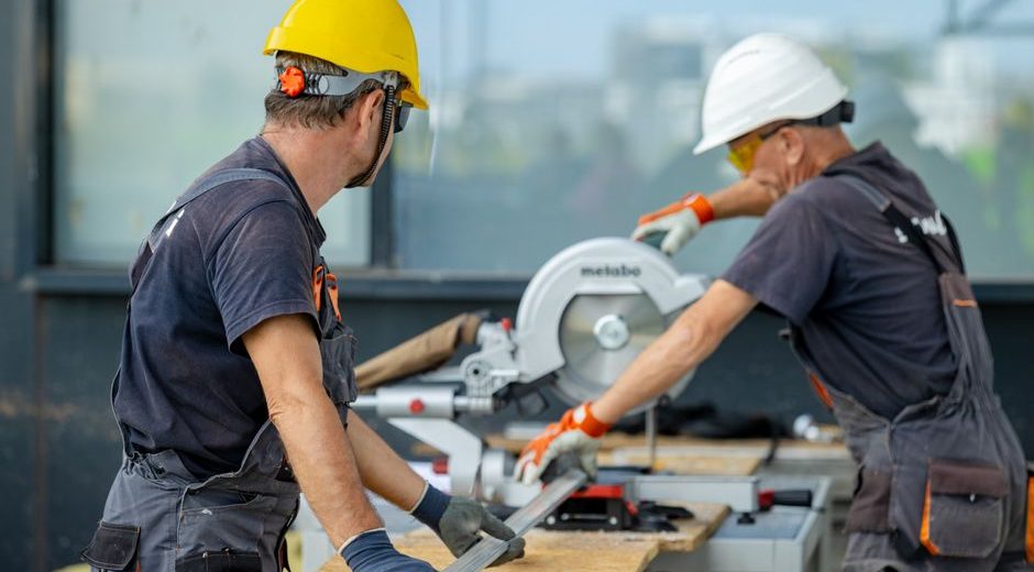 Two construction workers cutting a metal beam with a circular saw, wearing safety gear on site.